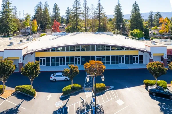 Aerial view of a commercial building with a curved white roof, parking lot, and tall evergreens with fall foliage.