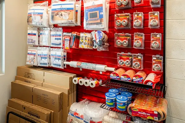 Red slatwall display of moving supplies including tape, locks, and stretch wrap, with stacked cardboard boxes below.