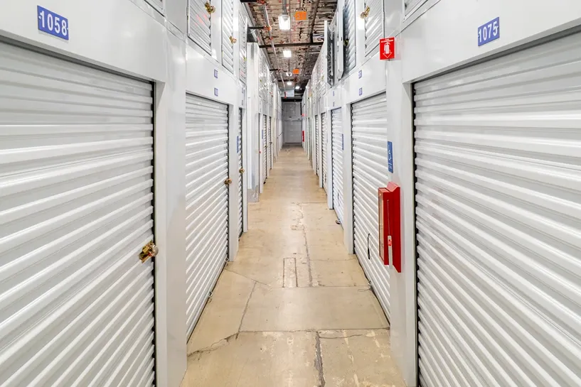 Indoor self-storage hallway with rows of numbered white roll-up doors, concrete floor, and a mounted fire extinguisher.